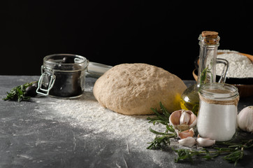 Ingredients for Focaccia: dough, rosemary, sea salt, olives, garlic, olive oil on a black wooden table.