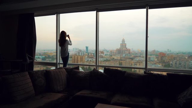 Young beautiful woman or tourist taking photograph with mobile cellphone smartphone device of skyline in Moscow Russia view from her apartment. Girl to share pic on social media photo site.