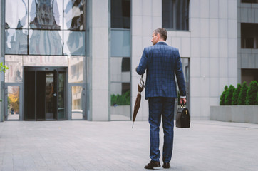 Stylish businessman in a trendy checkered jacket with an izont briefcase enters the front doors of a concrete gray business center.