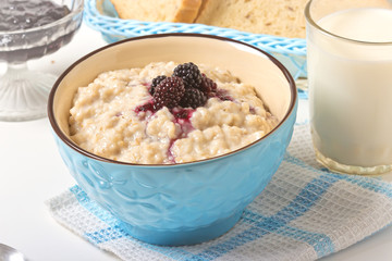 Healthy breakfast with a glass of milk, homemade oatmeal with blackberries in a blue bowl and fresh cereal bread on a light background