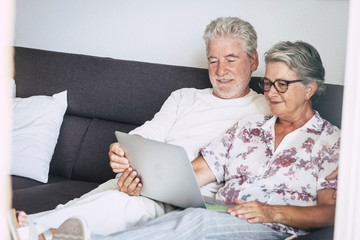 Beautiful caucasian aged senior couple at home watching movie or searching on the web sitting on the sofa with a laptop on the legs - indoor home technology concept for old people