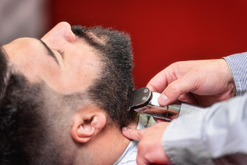 Barber shaving the beard of a handsome bearded man with an electric razor at the barber shop .