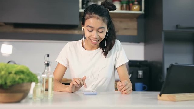 Happy, smiling young woman swiping and texting smartphone screen and listening to music in the headphones at the kitchen.