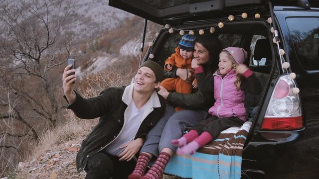 Family  sitting in the open trunk of a black car with kids and make a photo on the phone or selfie against the backdrop of autumn mountains