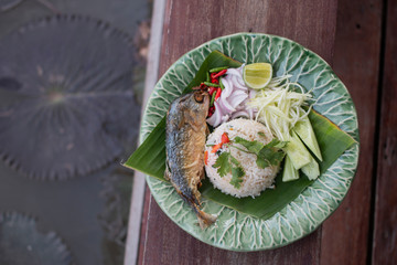 Tom Yum Fried Rice with Fried mackerel, chili, cucumber, shallot, sliced green mango, and lime serves on a beautiful plate.