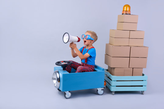 Smiling boy driving blue, fanny car and wagon with small boxes.Happy child with paper boxes on grey background. Studio shot. Delivery, shipping,  transport and supplier concept. 