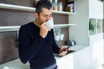 Handsome young man using his mobile phone while drinking coffee in the kitchen at home.