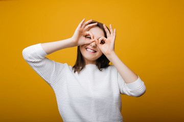 Portrait of happy caucasian girl looking through hands having fun against yellow background.