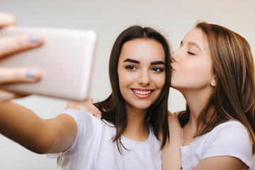 Two beautiful girlfriends doing selfie smiling while one is kissing another on the head in front of a white background.