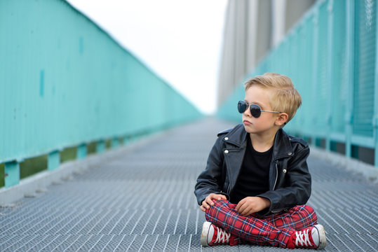 A Child rebel. A boy, 5 years old, fashionably dressed, poses like a model on the bridge. He is wearing a black leather jacket, red pants and sunglasses.