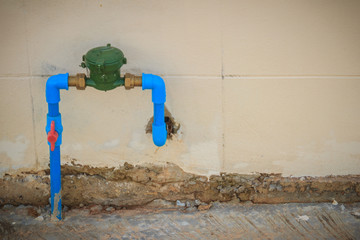 Vintage water meter installed with bronze joint, PVC elbow connector, blue PVC pipe and red water valve on the grungy wall.