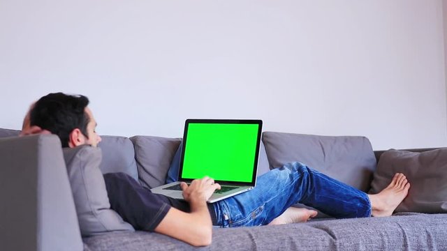 Young man slying down on sofa and using laptop computer. Candid view