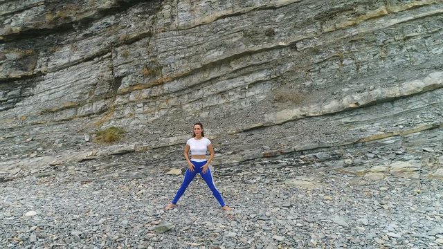 Woman is doing squats the bodyflex during breathing exercises on the rock background. Aerial beautiful footage view.