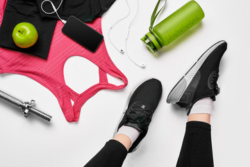 Girls legs wearing black trainers sitting on a mat surrounded by mobile phone, earphones, towel and...
