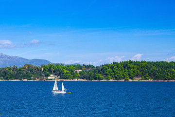 Obraz premium Wonderful romantic summertime panoramic seascape. Sailing yacht with white sails in to the crystal clear azure sea. Small green island against coastline slopes.