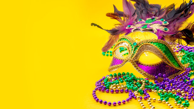 Happy Mardi Gras and Fat Tuesday carnival concept with close up on a face mask full of color, feathers and texture and golden, green and purple beads isolated on yellow background with copy space