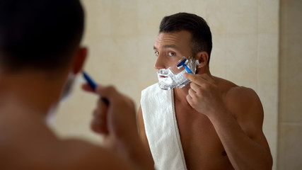 Male with foam on face using manual razor to trim and shave beard in bathroom