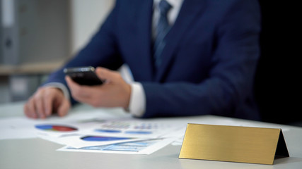 Business man in suit using phone, annual report on table, golden blank nameplate