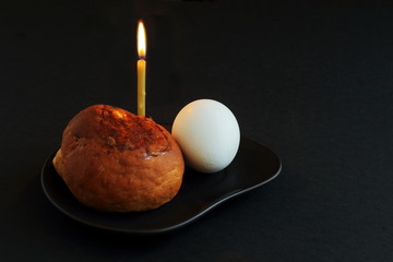 Delicious, fragrant bread, white egg and burning wax candle on black clay plate on dark background. Easter concept.