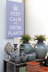 Stone birds, ceramic vases, flower pots standing on a wooden table and hanging picture "Keep calm and grow plants" in a flower shop