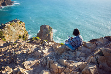 Young woman sitting is using smartphone on mountain cliff edge on ocean sunny natural background. Traveler having social communication technology.