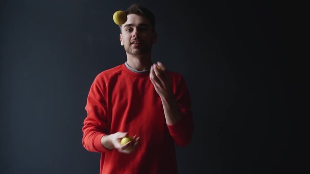 Young man in coral red sweater motivationally juggling lemons, isolated on dark background - slow motion