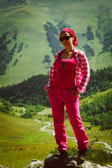 girl in pink pants and plaid shirt standing on a rock on a rock hands in pockets on a mountain background