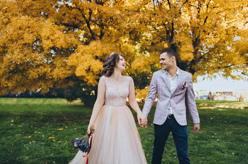 Newlyweds in love walk in the fall in the park with yellow foliage. Stylish groom in a plaid jacket leads the hand of a cute bride in a beige dress with a crown. Wedding photography. Princess Bride.