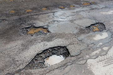 Potholes on a paved road. The road is in very poor condition. There is dirty water in the potholes.