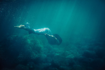 brunette girl in long blue dress dives underwater in the sea - obrazy, fototapety, plakaty
