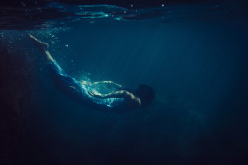 brunette girl in long blue dress dives underwater in the ocean