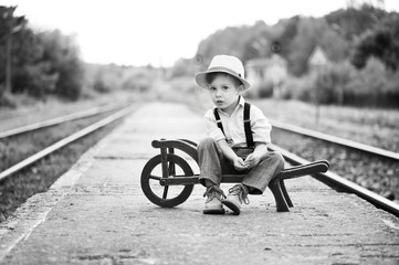 Monochrome portrait of cute boy wearing in retro style sitting on railway station and is waiting...