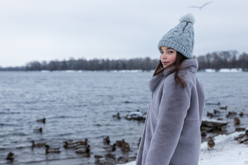 background. a beautiful girl in a gray hat and a fur coat, in winter on the banks of a river / lake / sea / ocean feeds ducks.