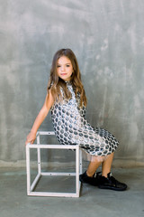 Full length of cheeful lovely little girl in grey fashion dress sitting on the white cube