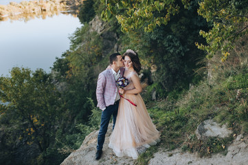 Autumn portrait of beautiful newlyweds, on a background of hills and rocks with greens. Wedding photography. Stylish groom hugs and kisses a young bride in a beige dress with a crown.