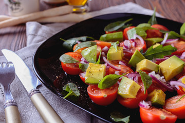 Colorful and tasty salad with avocado, cherry tomatoes and mozzarella on a black dish next to a fork and knife served to eat