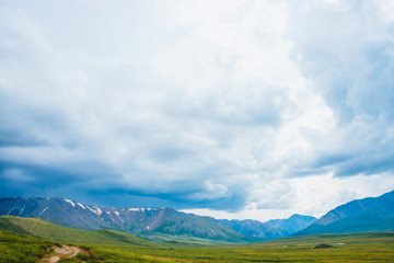Fototapeta premium Spectacular view of distant giant mountains. Footpath through valley in highlands. Hiking path. Wonderful huge mountain range under cloudy sky. Amazing dramatic green landscape of majestic nature.
