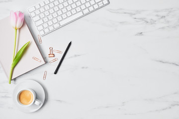 Office desk with computer, pink tulip, coffee cup and pink diary on white marble background. Flat lay. Copy space for text. Top view.