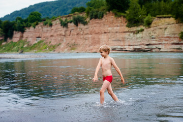 Blonde smiling boy in red swimsuit running in the mountain river