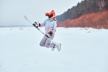 Cheerful woman wearing warming sportswear holding a snowboard and jumping on a snowy beach