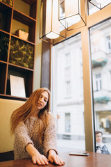 Portrait of young pretty caucasian hipster blondie girl sitting on a sofa, relaxing, dreaming, near big window. Lifestyle, beauty, youth concept