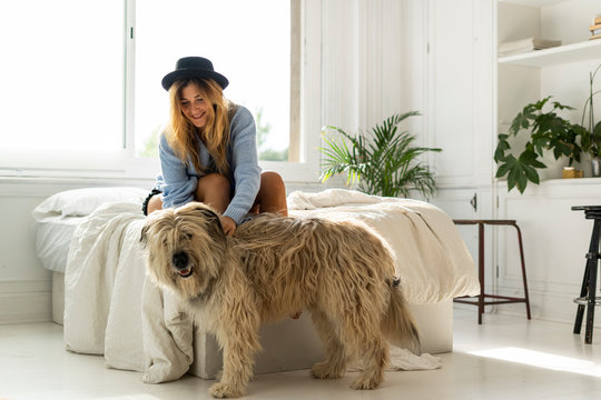 Smiling young woman sitting on bed stroking a dog