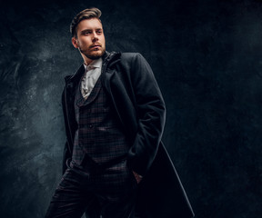 A stylish young man dressed in an elegant suit and coat posing with a hand in a pocket against a...