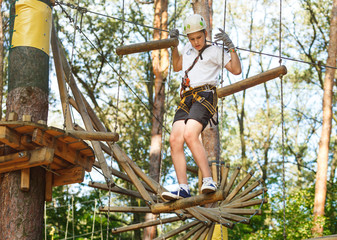 Cheerful cute young boy in white t shirt and white helmet in adventure rope park at sunny summer day. Active lifestyle, sport, holidays for children