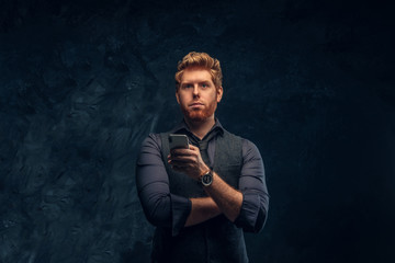 Portrait of a redhead man in formal wear holding a smartphone in studio against a dark textured wall