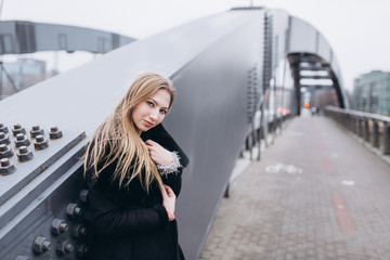 Fototapeta premium Young blondie sexy sensual girl go across the bridge in black coat. Outdoor portrait. Casual, lifestyle concept