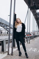 Fototapeta na wymiar Young blondie sexy sensual girl go across the bridge in black coat. Outdoor portrait. Casual, lifestyle concept