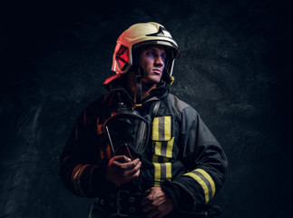 Studio portrait in a dark studio against a textured wall. Brutal firefighter in uniform and safety helmet holding an oxygen mask and looking sideways with a confident look.