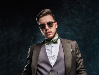Fototapeta premium A stylish man in sunglasses dressed in an elegant suit against a dark textured wall