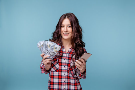 Young girl isolated over blue background. Looking camera showing display of mobile phone holding money